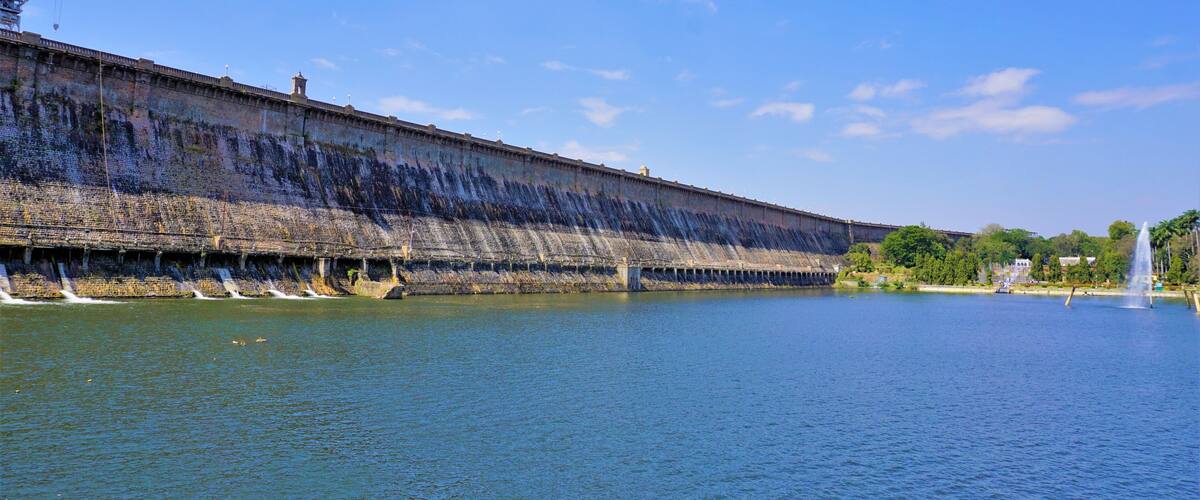 A large dam with water flowing over its spillway, a clear blue sky, and a fountain in the foreground.