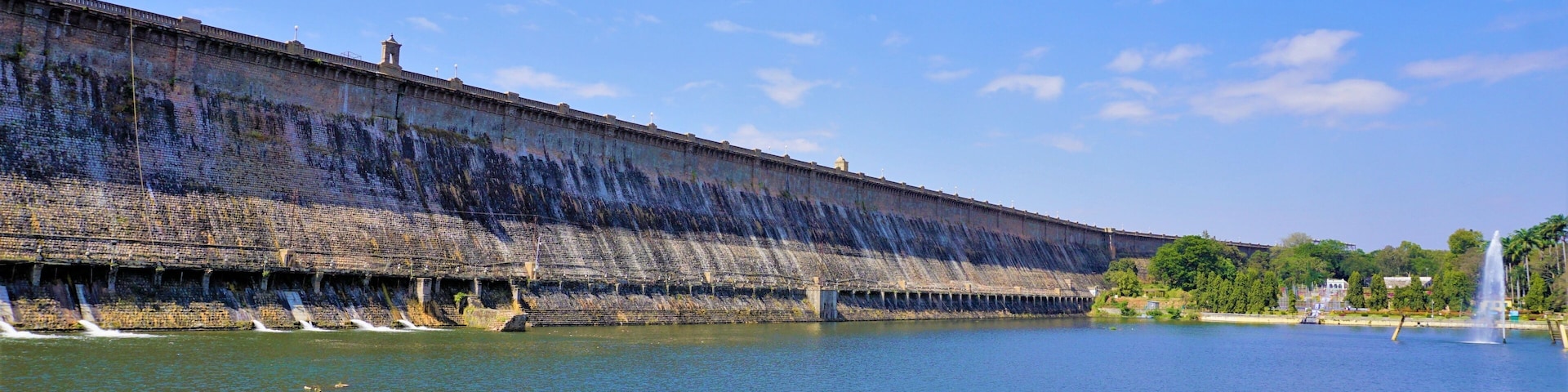 A large dam with water flowing over its spillway, a clear blue sky, and a fountain in the foreground.