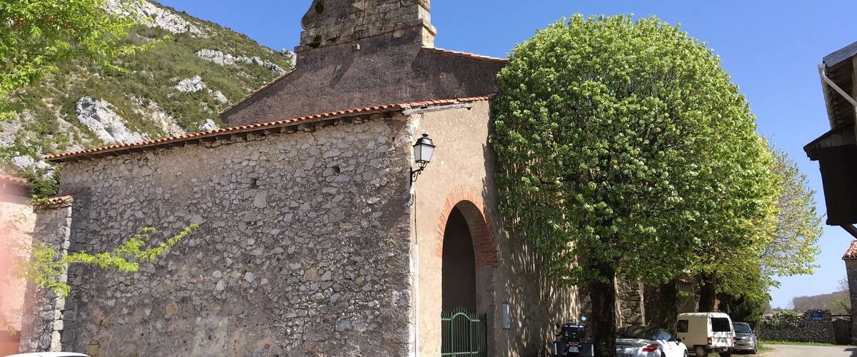 A stone church with a bell tower, parked cars, and a tree in a sunny village setting.