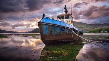 A blue and rusted boat tied to a dock with a cloudy sky and mountains in the background.