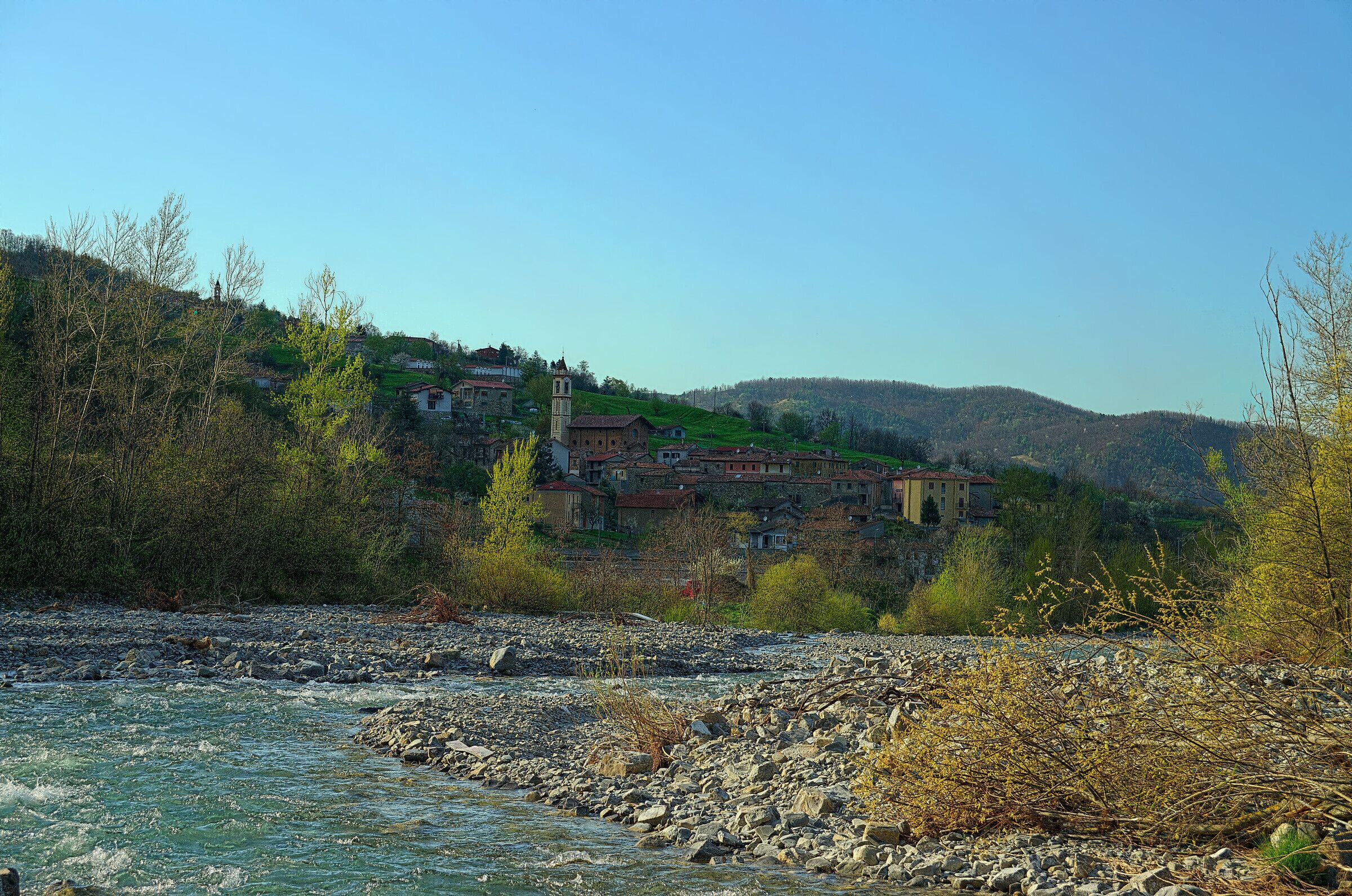 A river with rocks, a village with buildings and a tower, and hills in the background.