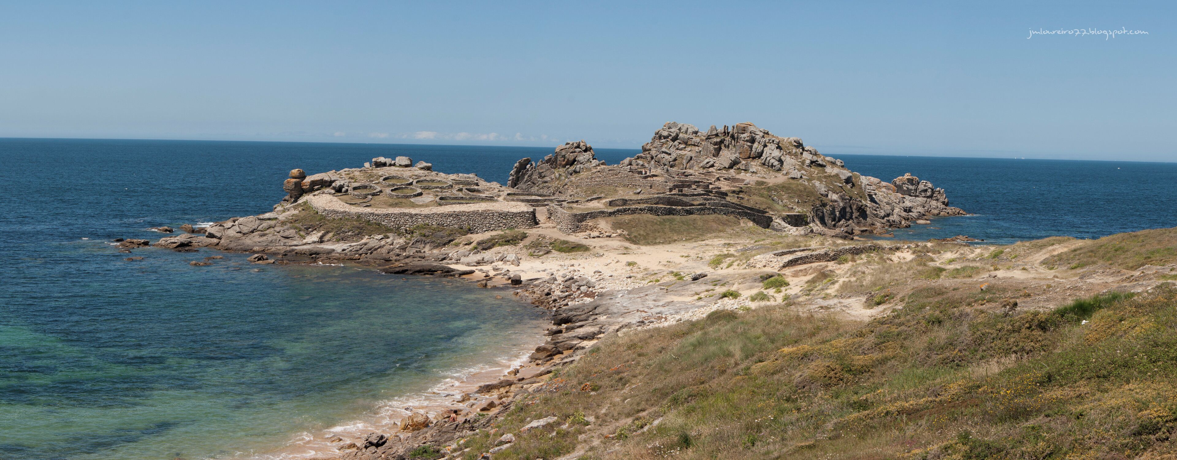 A rocky coastal landscape with clear blue waters and a small island with ancient ruins.