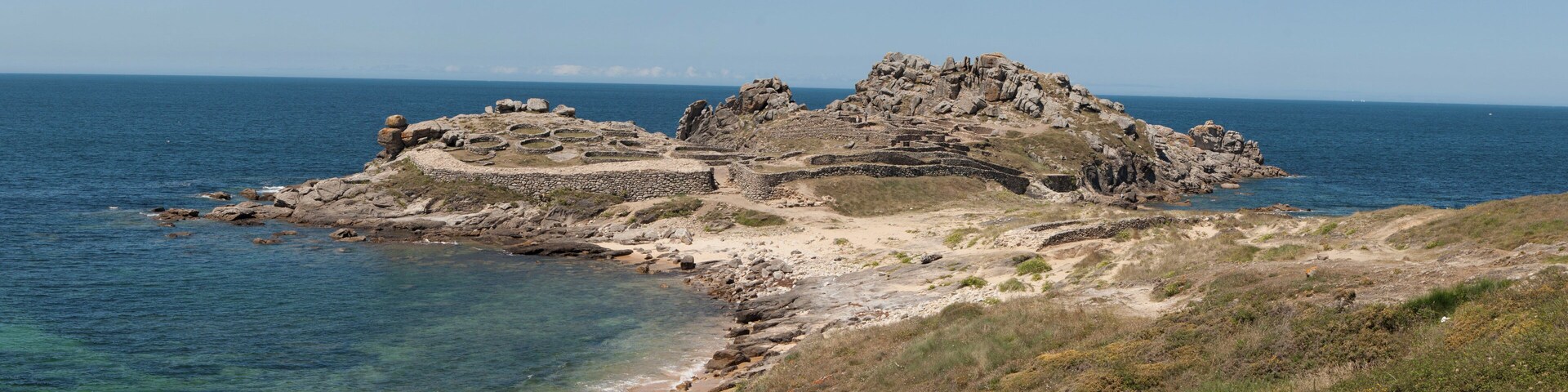 A rocky coastal landscape with clear blue waters and a small island with ancient ruins.