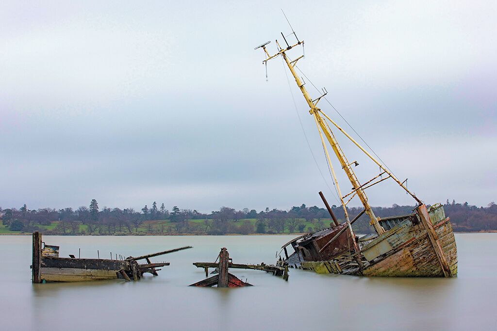 A derelict shipwreck in a calm lake with a forested shoreline in the background.
