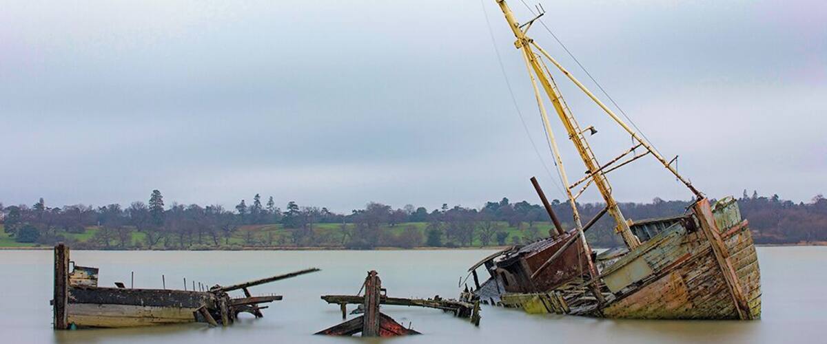 A derelict shipwreck in a calm lake with a forested shoreline in the background.