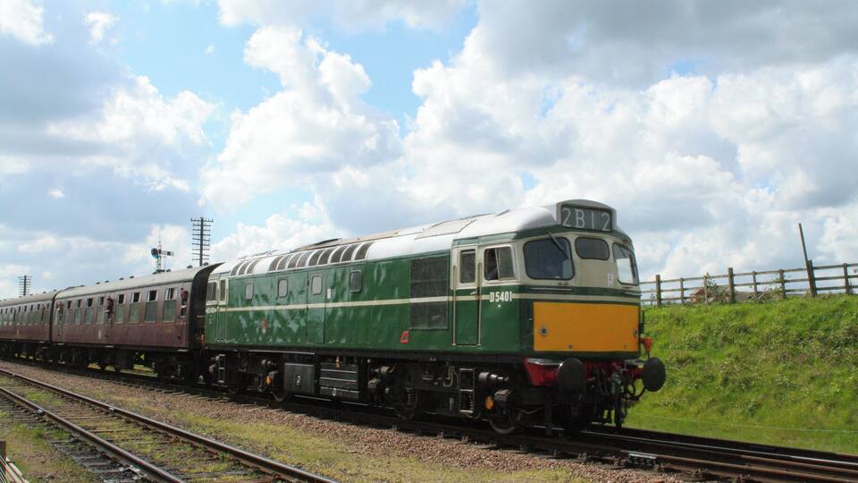A green and white train with the number 2B12 on the front, traveling on tracks with a grassy embankment and a wooden fence in the background.