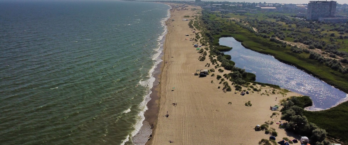 A coastal beach with a river running parallel to the shore, surrounded by greenery and urban buildings.