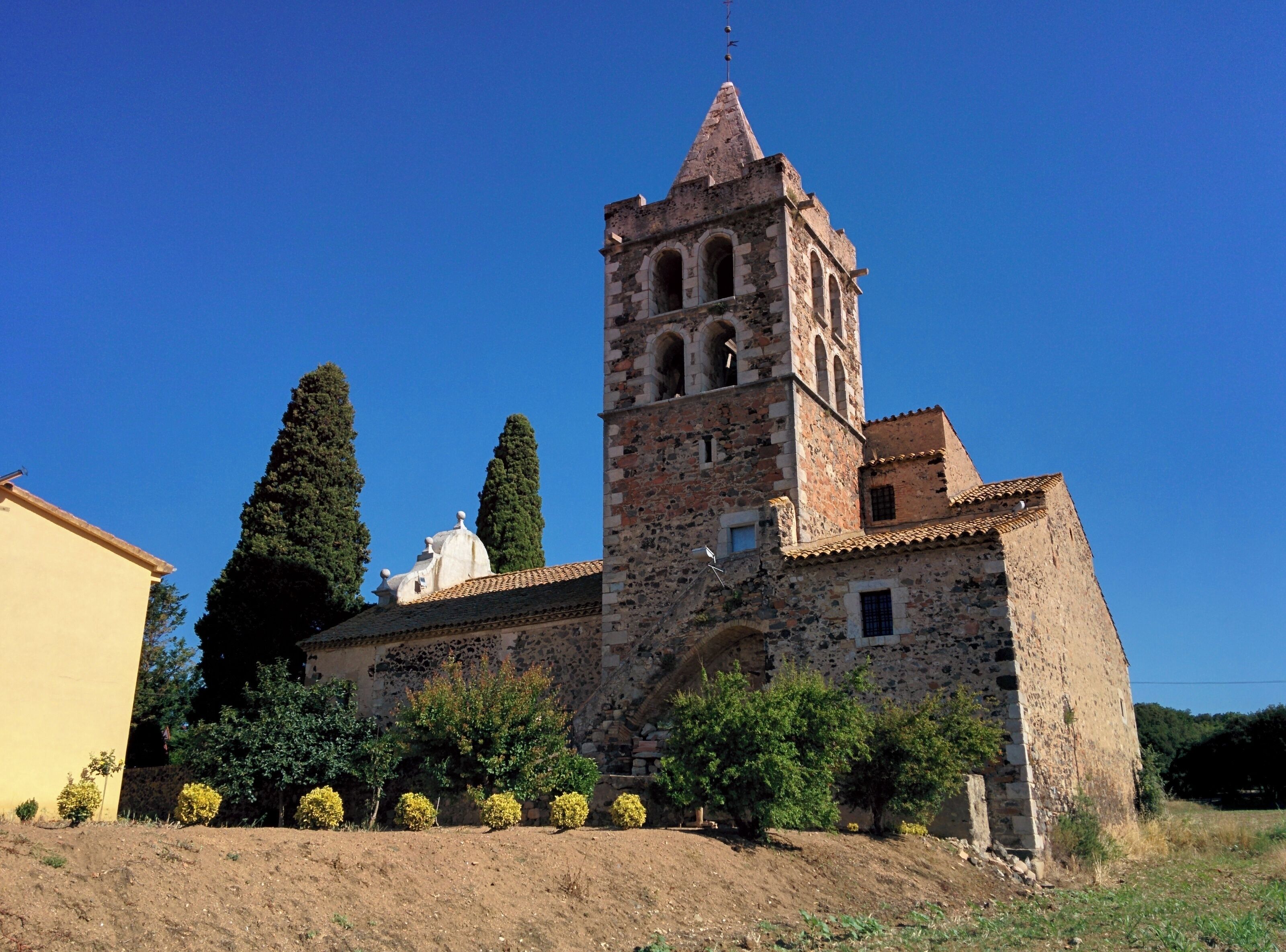 A historic stone church with a bell tower, surrounded by greenery and a clear blue sky.