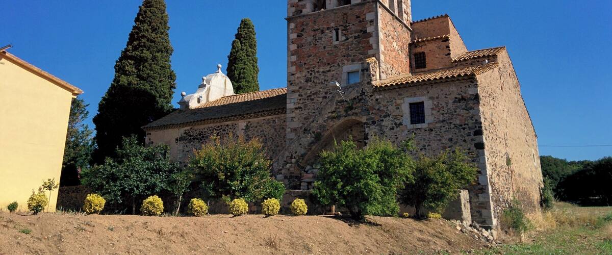 A historic stone church with a bell tower, surrounded by greenery and a clear blue sky.
