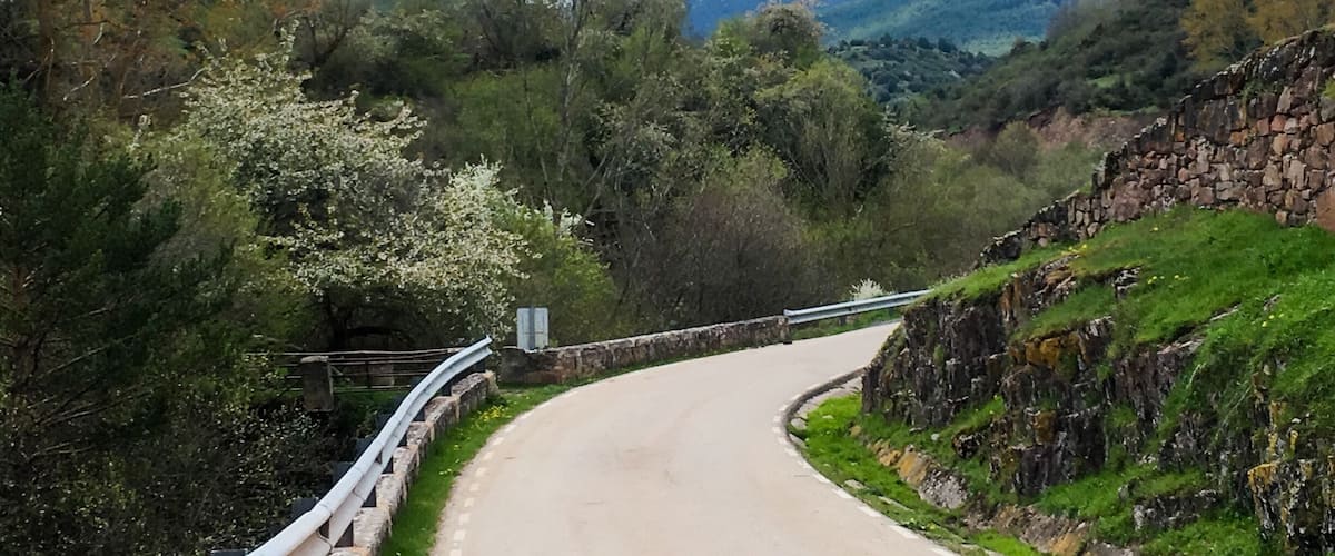 A winding mountain road with a stone wall on one side and a snow-capped mountain in the background.
