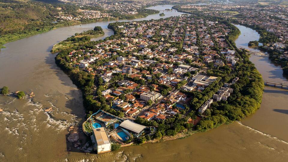 An aerial view of a riverine town with dense housing, a prominent swimming pool complex, and surrounding greenery.