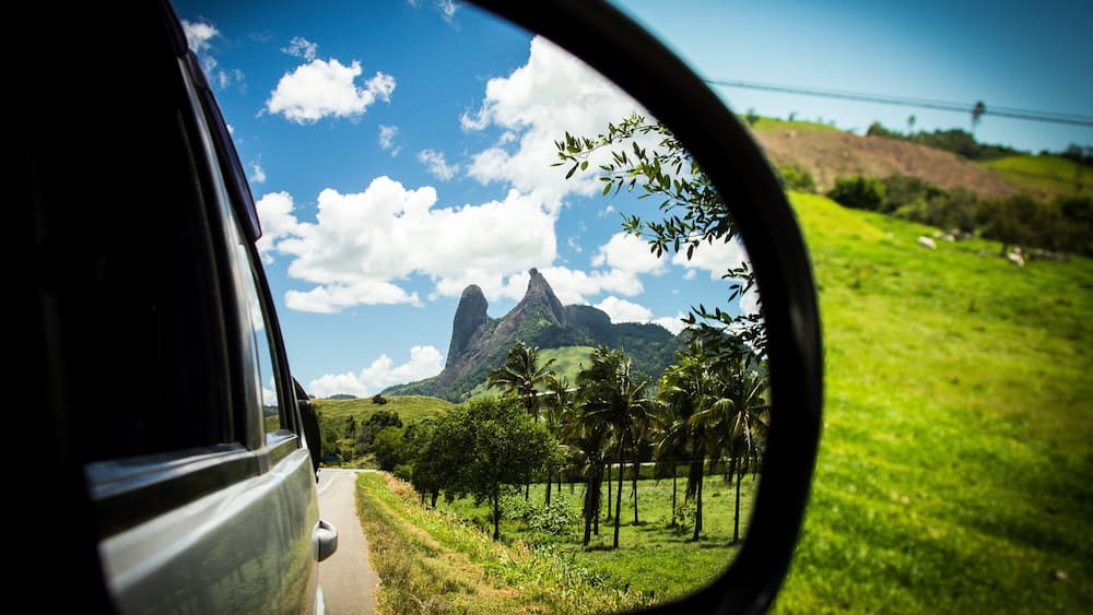 A car's side mirror reflecting a road, green grass, palm trees, and a mountain with a peak.