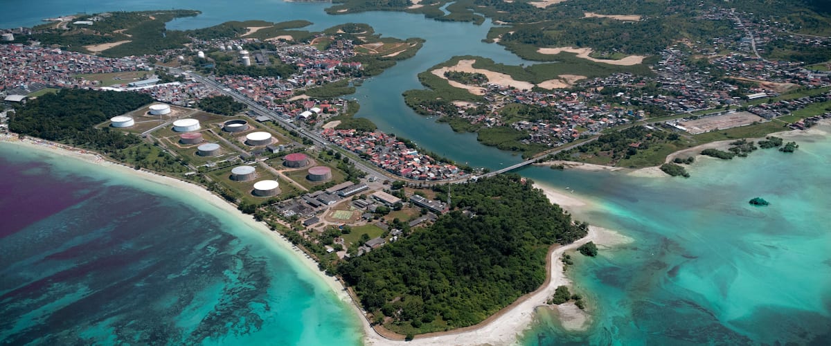 An aerial view of a coastal town with a beach, industrial facilities, and a dense forest area.