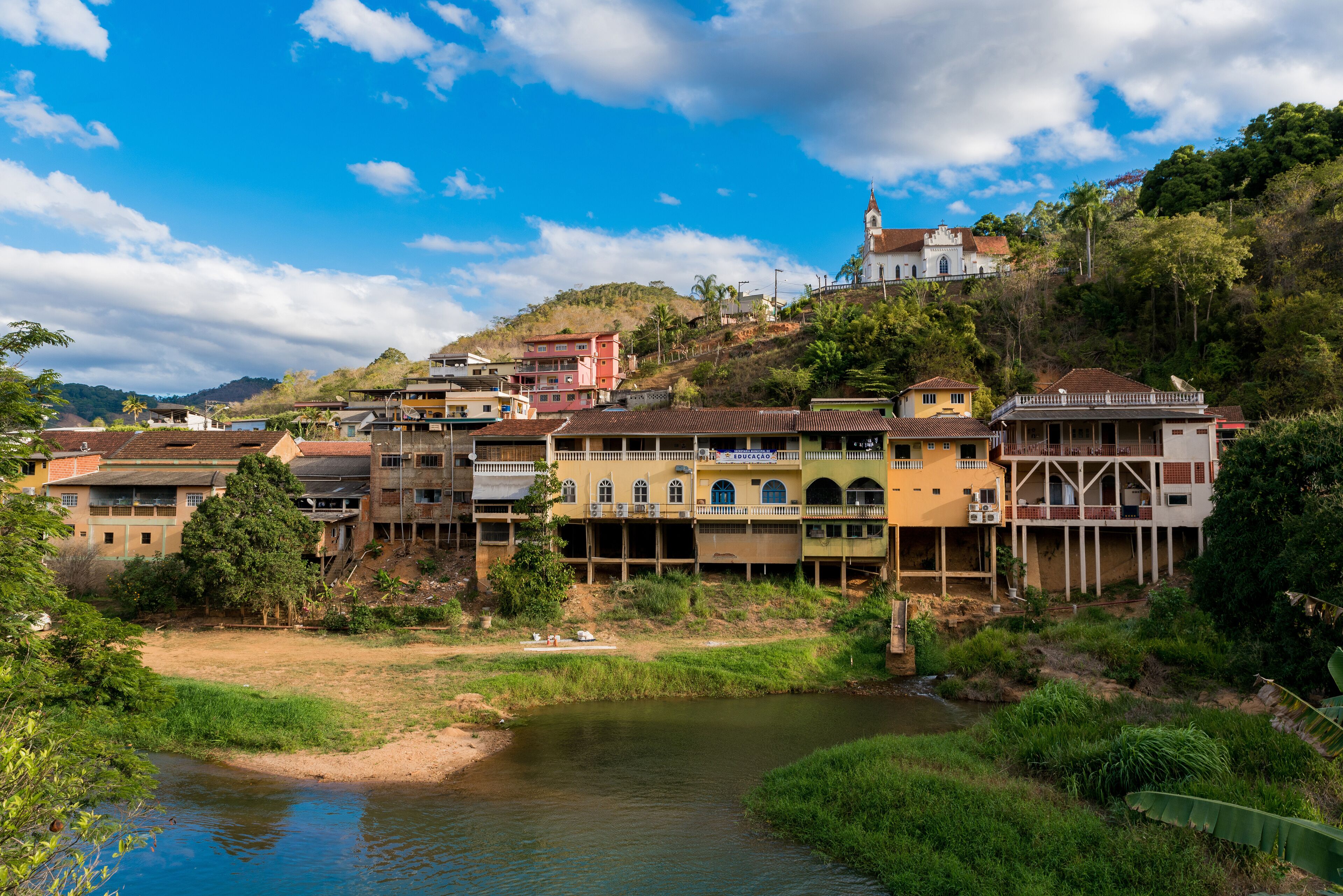 A riverside town with colorful buildings, a church on a hill, and lush greenery.