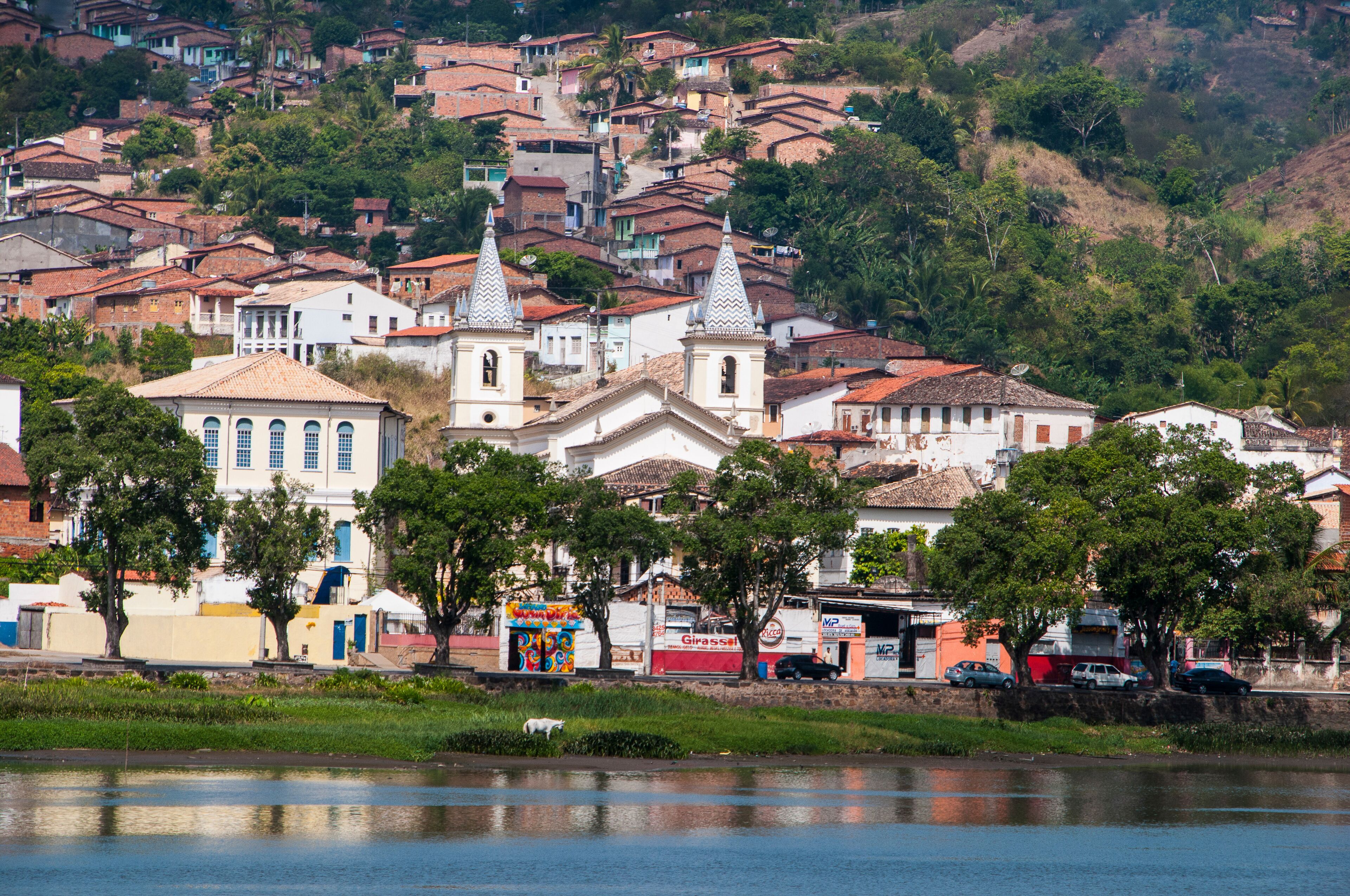 A hillside town with terracotta-roofed houses, a prominent church with two towers, and a river in the foreground.