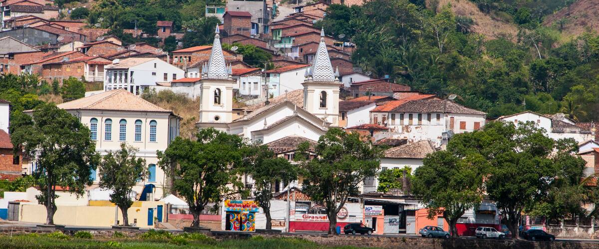 A hillside town with terracotta-roofed houses, a prominent church with two towers, and a river in the foreground.