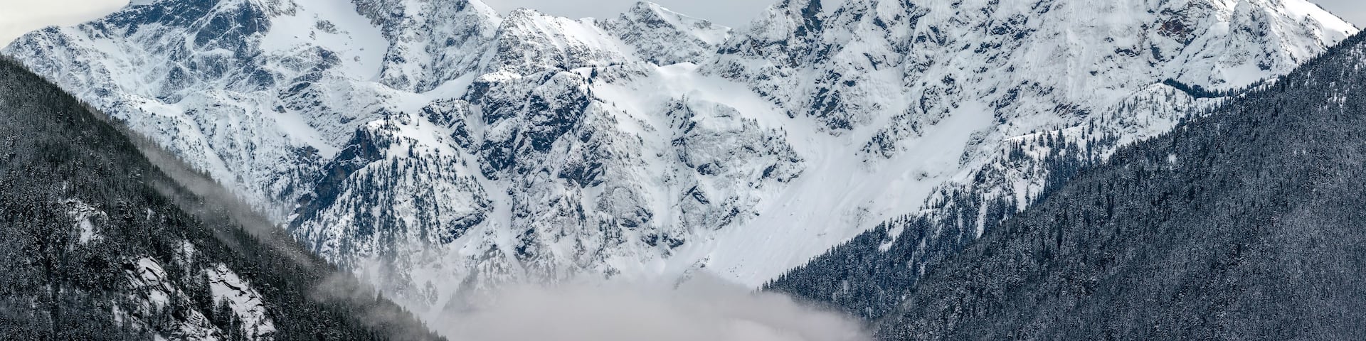 A snow-covered mountain range with sharp peaks and dense forests at the base.