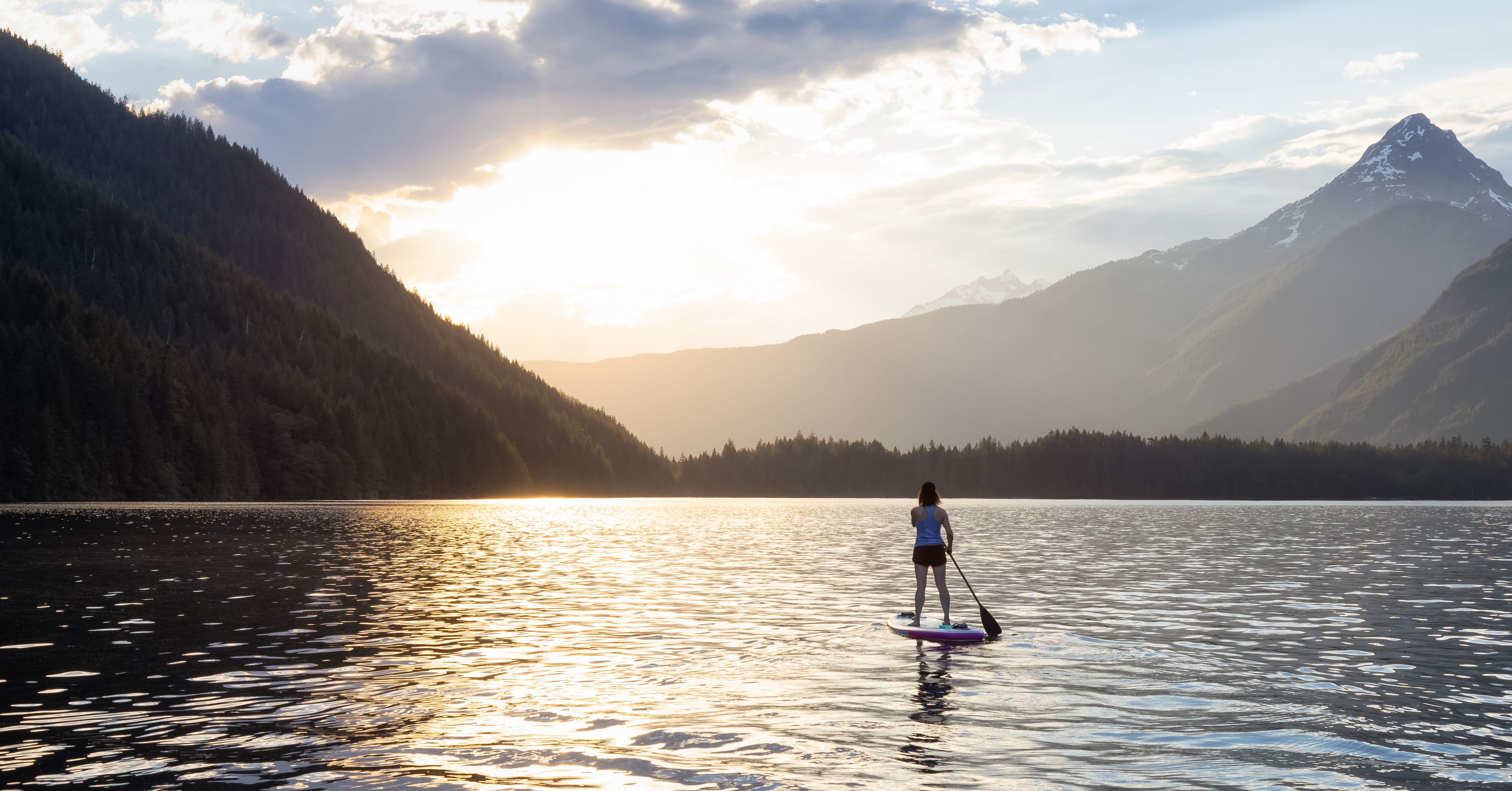 Une personne fait de la planche à pagaie sur un lac calme, avec des montagnes en arrière-plan.