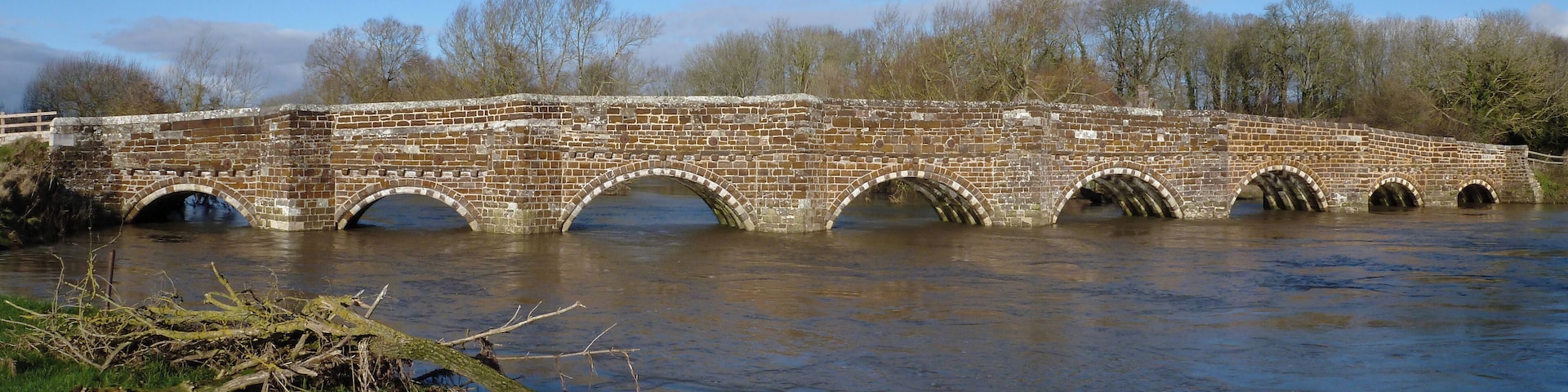 A stone bridge over a river with grassy banks and trees in the background.