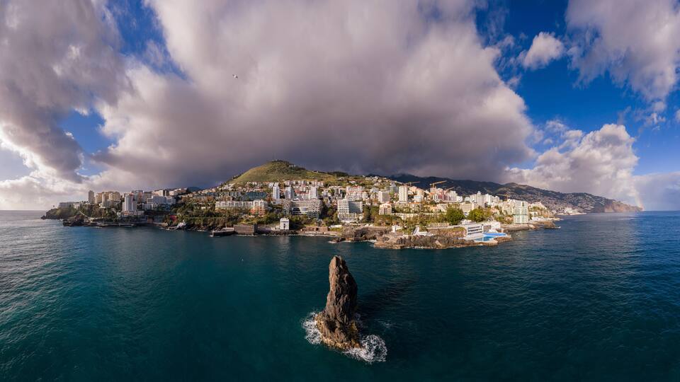 A coastal cityscape with a prominent hill, a rocky outcrop in the water, and a clear sky.