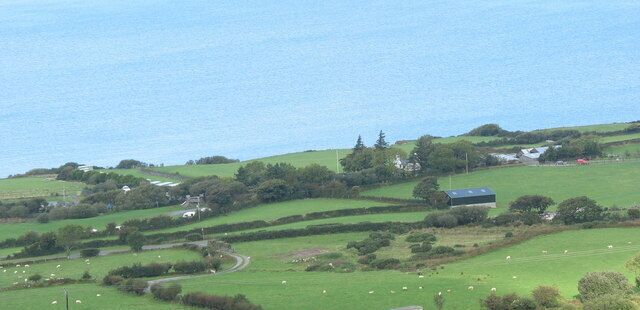 A coastal landscape with rolling hills, green fields, scattered sheep, and a few buildings.
