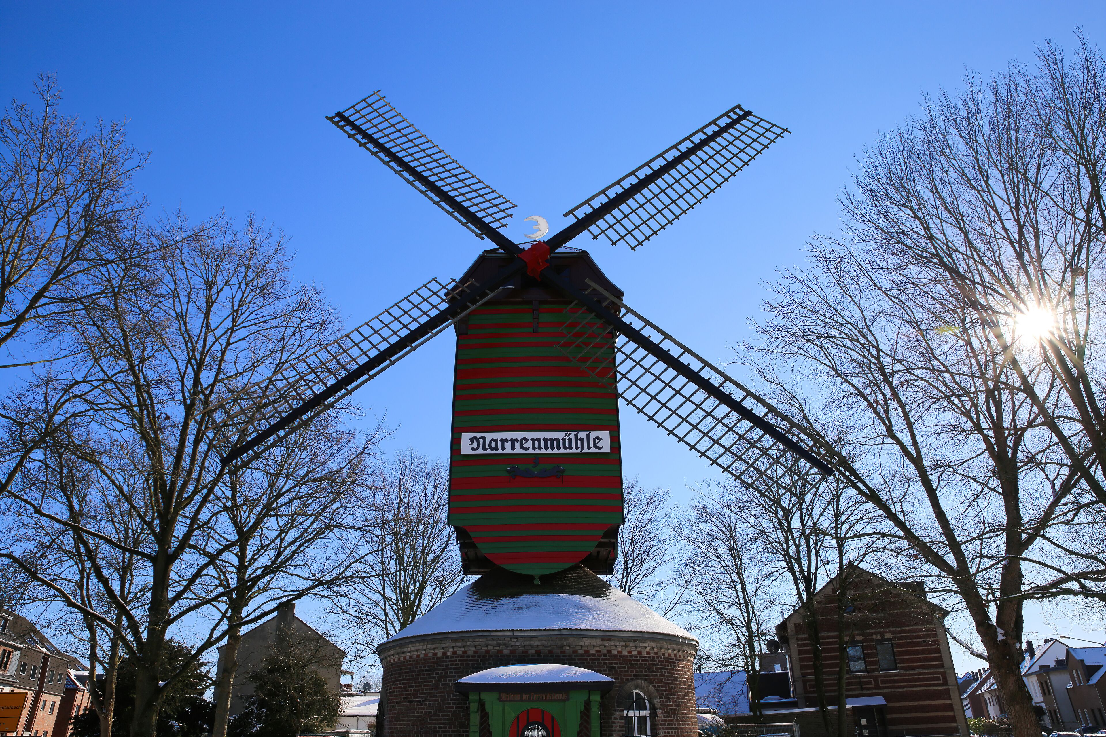 Viersen (Dulken), Germany - February 9. 2021: View over snow on red green colorfull striped wind mill (Narrenmuhle) with bare trees, sun lens flares