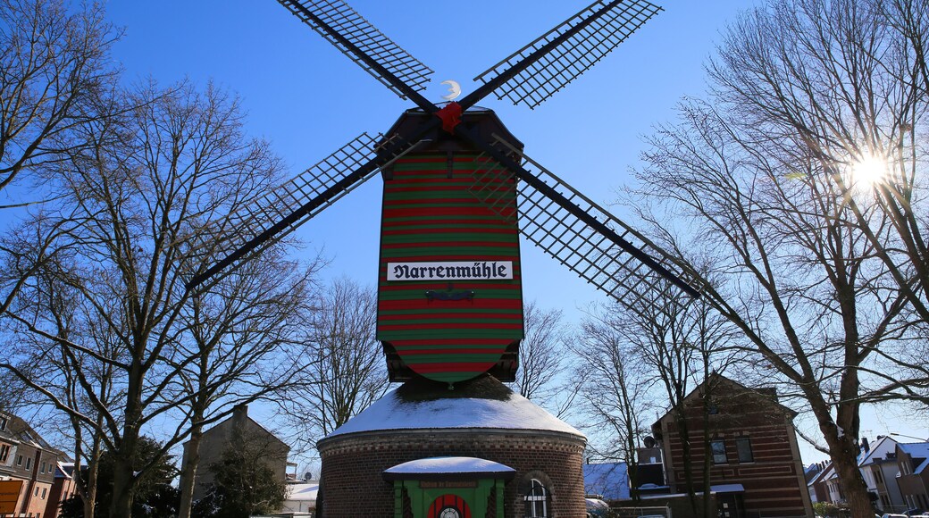 Viersen (Dulken), Germany - February 9. 2021: View over snow on red green colorfull striped wind mill (Narrenmuhle) with bare trees, sun lens flares