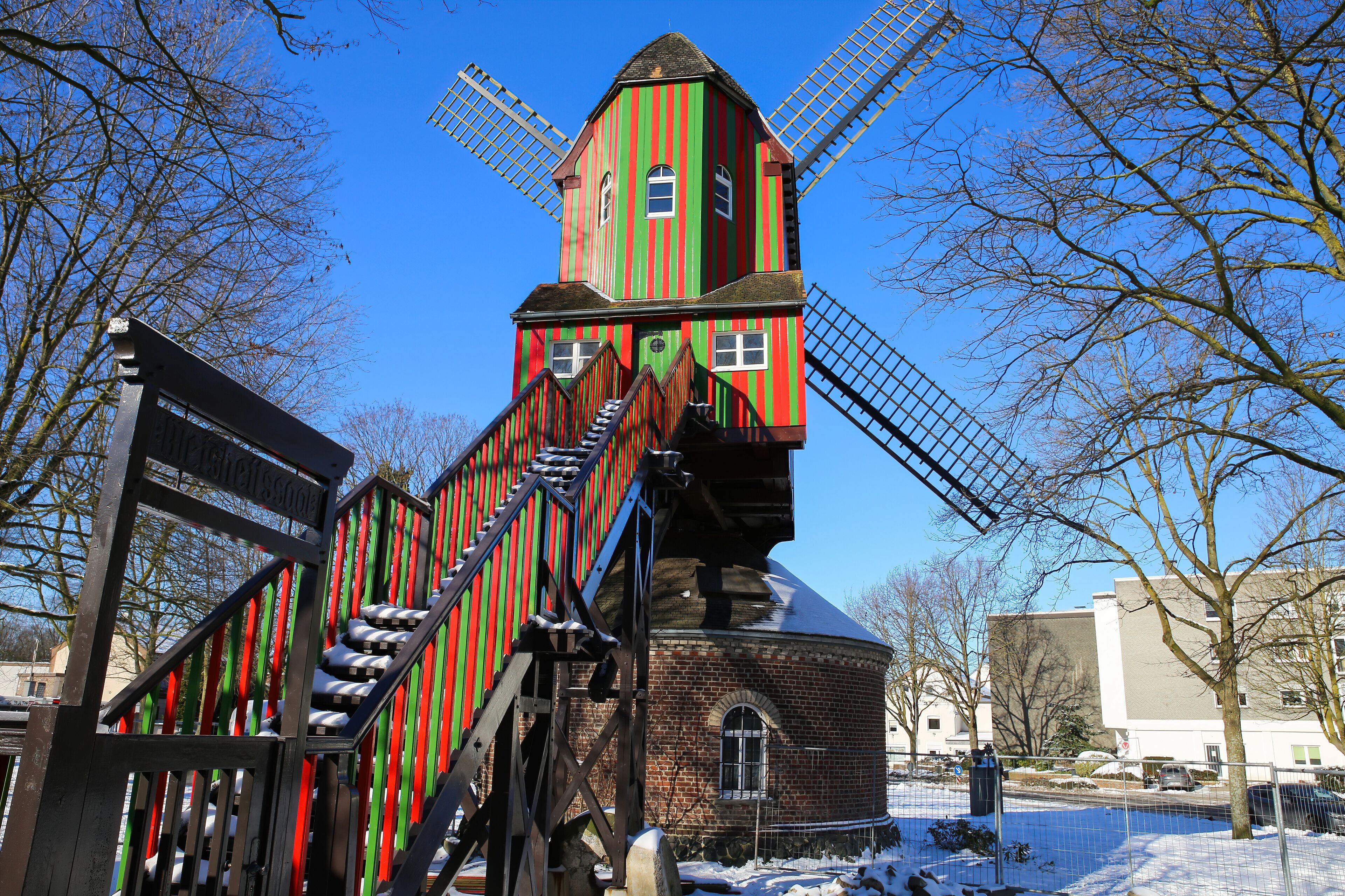 A colorful windmill with a red and green striped facade, a brick base, and a metal staircase leading up to the top.