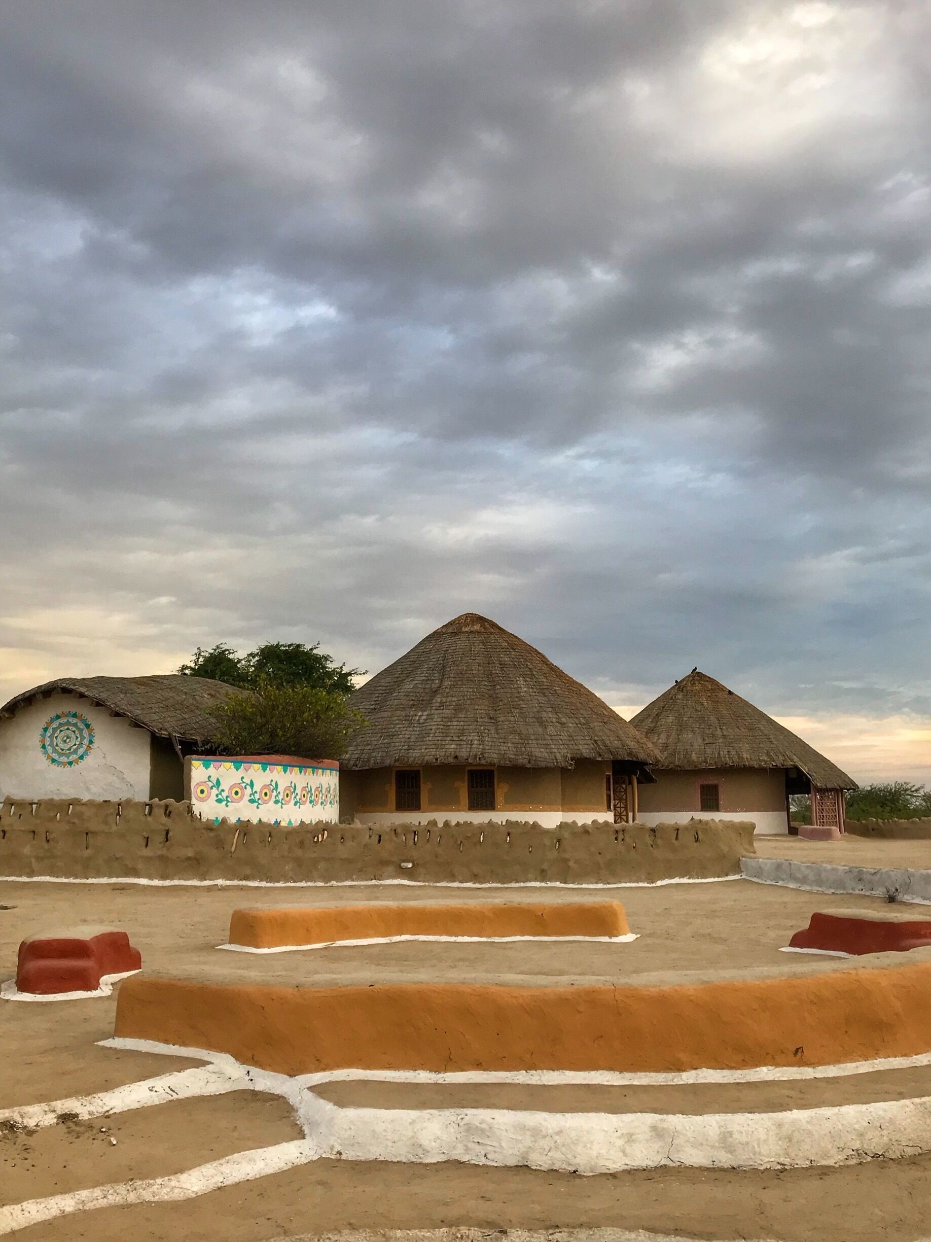 Traditional thatched-roof huts with colorful wall decorations, set against a cloudy sky.