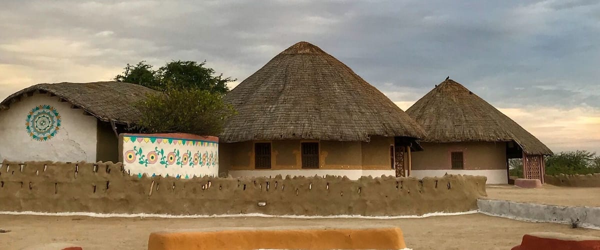 Traditional thatched-roof huts with colorful wall decorations, set against a cloudy sky.