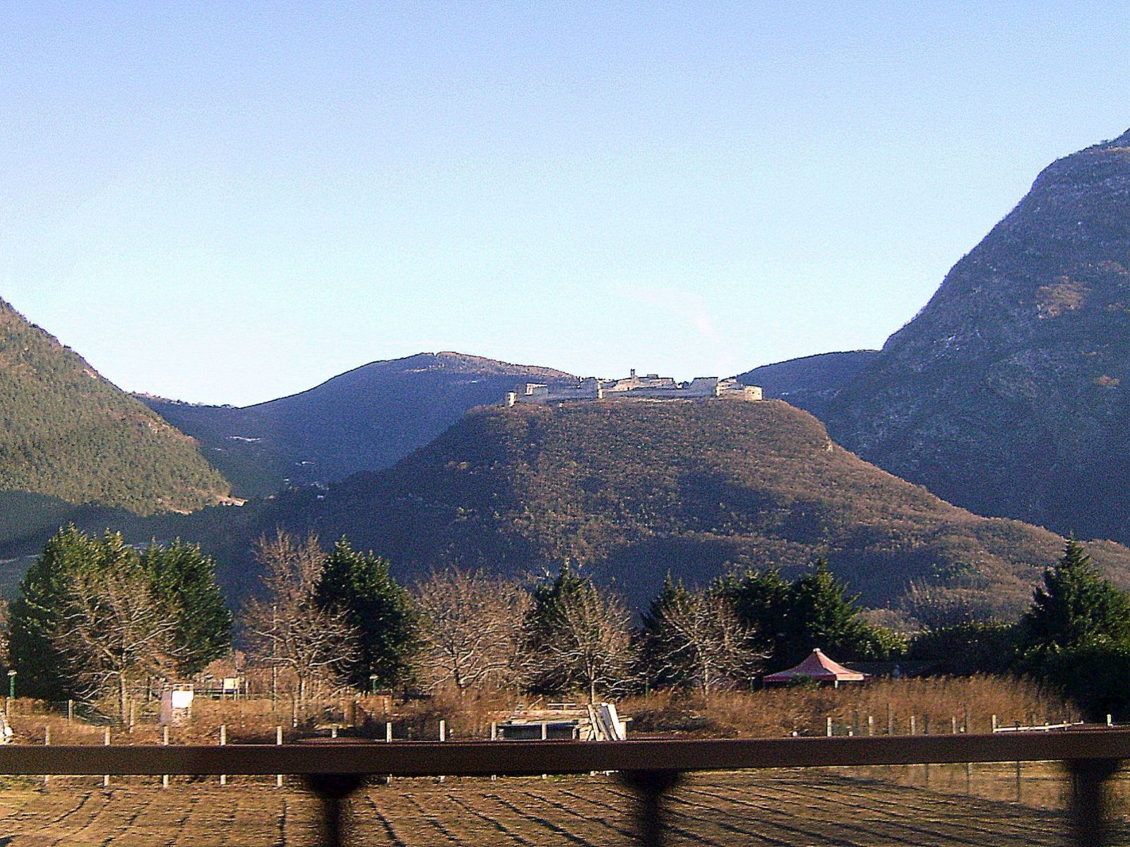 A mountain with a small settlement on top, surrounded by forested hills.