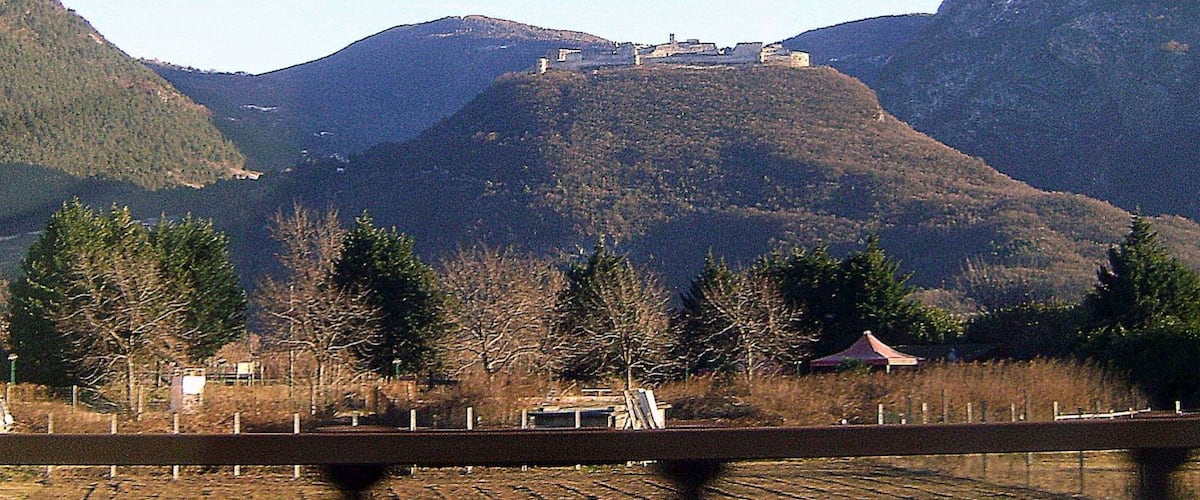 A mountain with a small settlement on top, surrounded by forested hills.