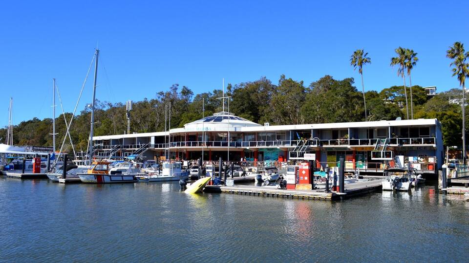A marina with boats docked, a large building with a sloped roof, and palm trees in the background.