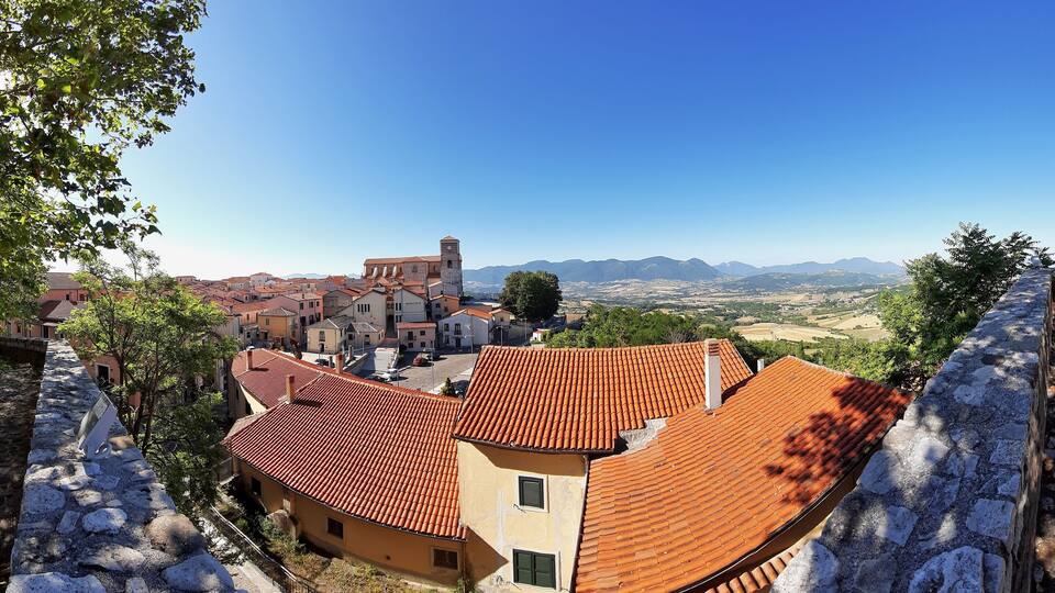 A town with red-tiled roofs, a prominent church with a bell tower, and a mountainous landscape in the background.