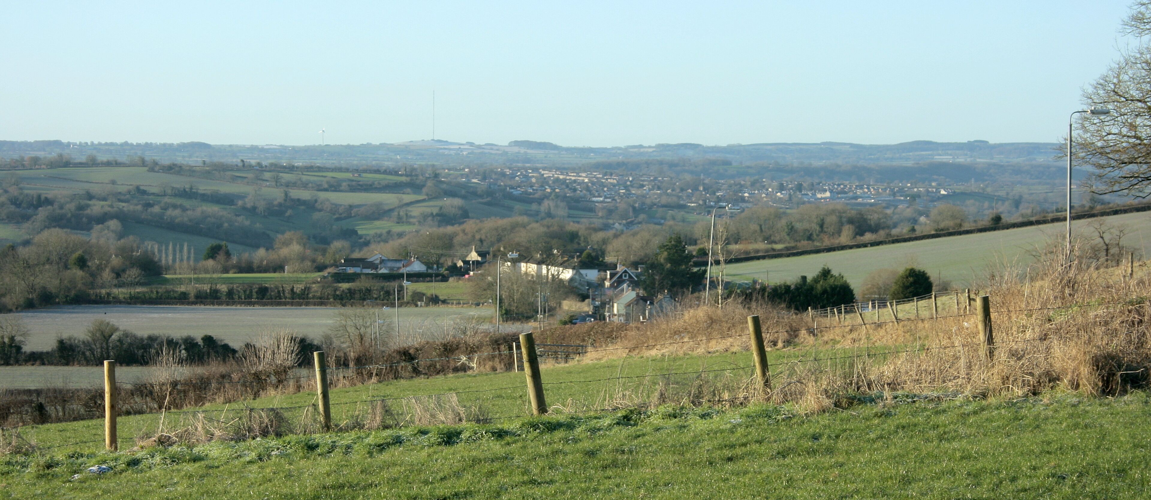 A rural landscape with rolling hills, a small village, and a distant wind turbine.