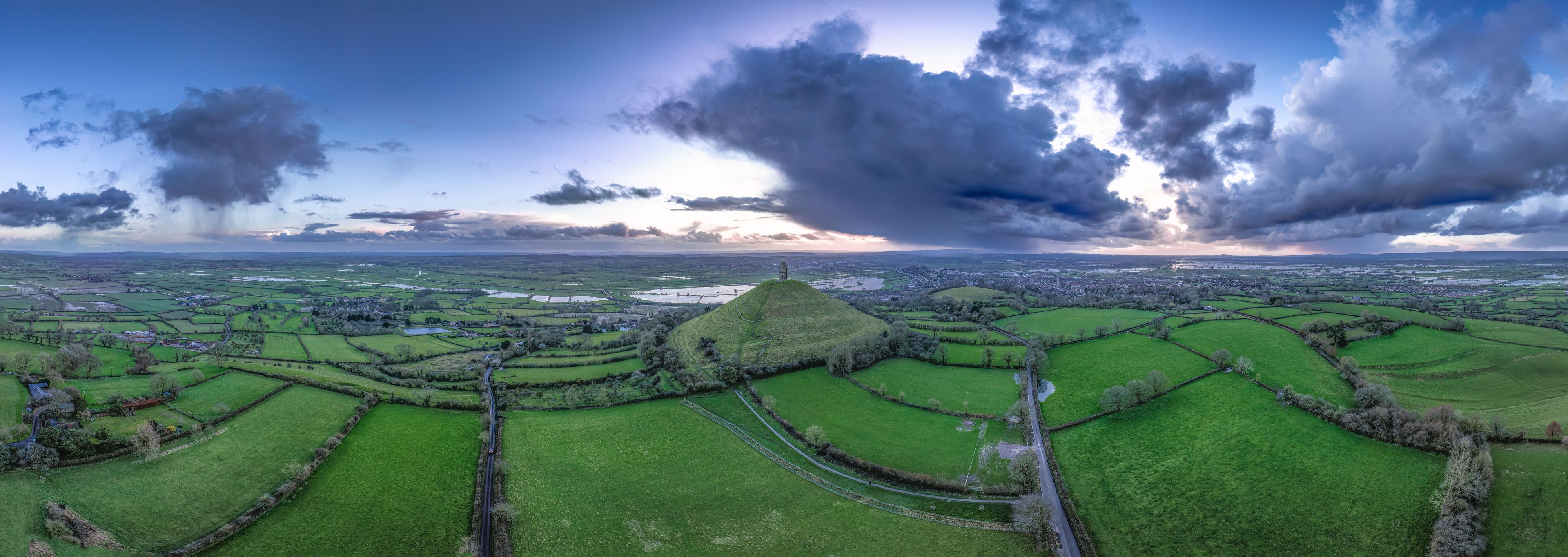 A rural landscape with rolling green fields, a winding road, and a prominent hill with a structure on top.
