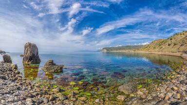 A rocky shoreline with clear water, a large rock formation, and a distant hill with sparse vegetation.