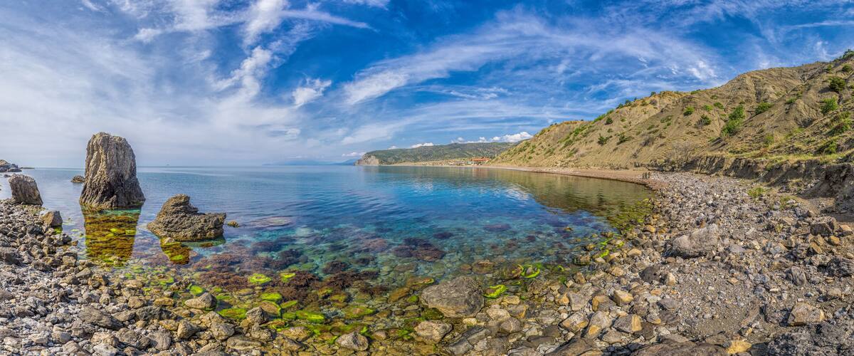 A rocky shoreline with clear water, a large rock formation, and a distant hill with sparse vegetation.