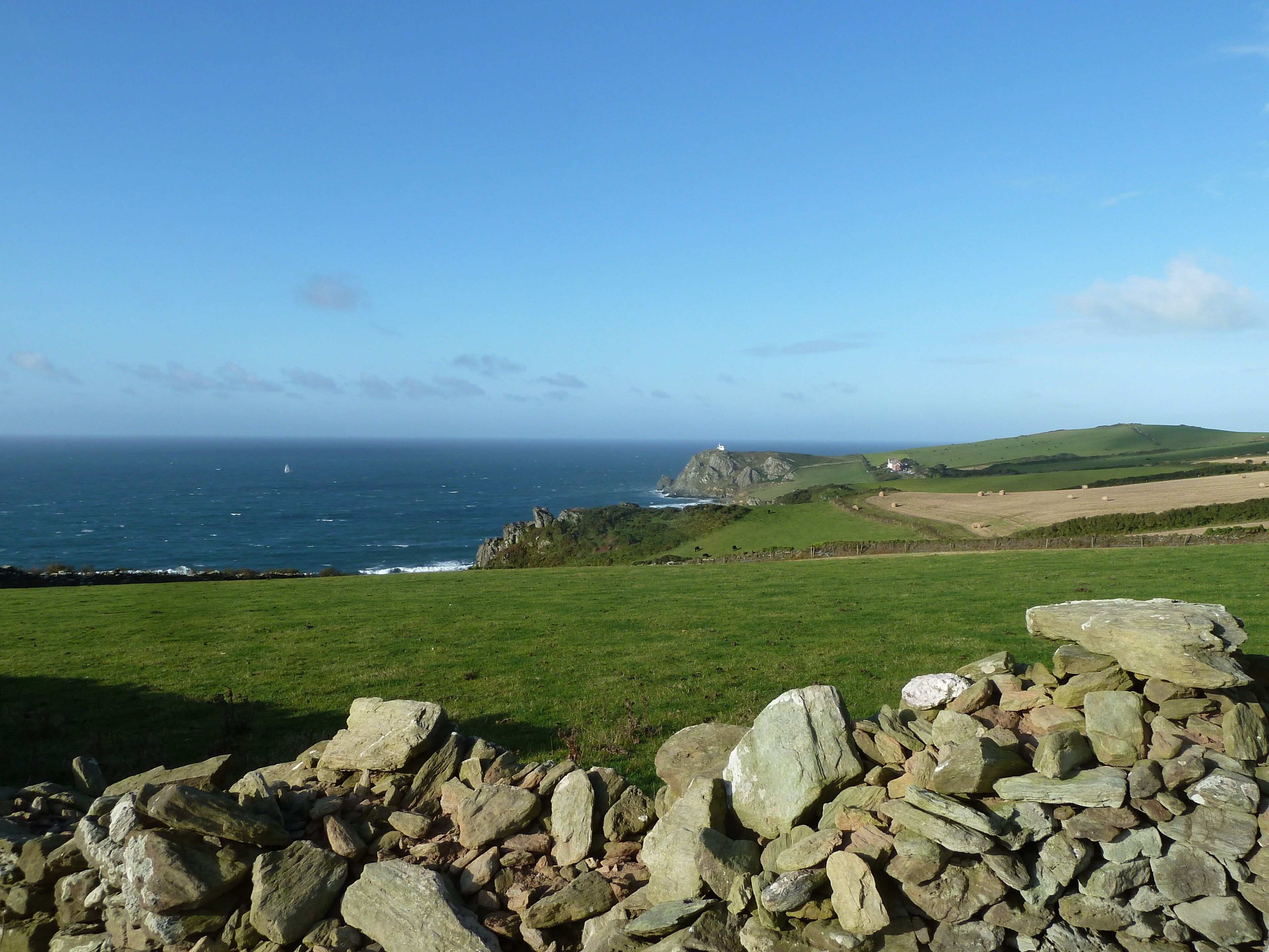 A coastal landscape with a stone wall in the foreground, a lighthouse on a rocky outcrop, and a grassy field with hay bales in the distance.