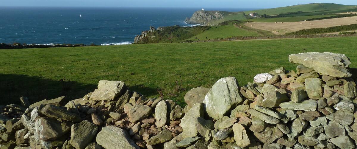 A coastal landscape with a stone wall in the foreground, a lighthouse on a rocky outcrop, and a grassy field with hay bales in the distance.