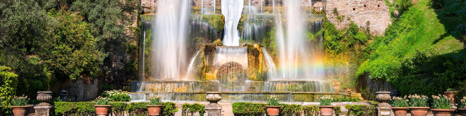 A historic fountain with multiple water jets, surrounded by lush greenery and classical architecture.