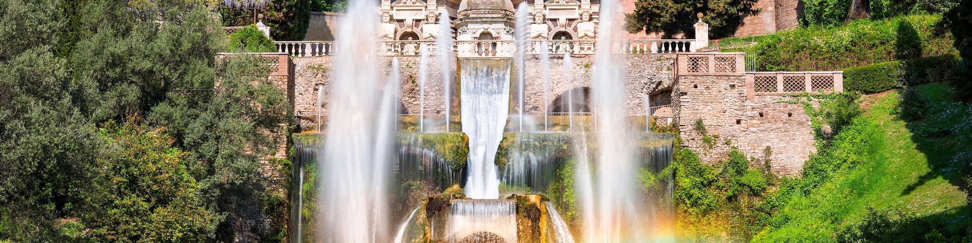 Una fuente histórica con múltiples chorros de agua, rodeada de exuberante vegetación y arquitectura clásica.