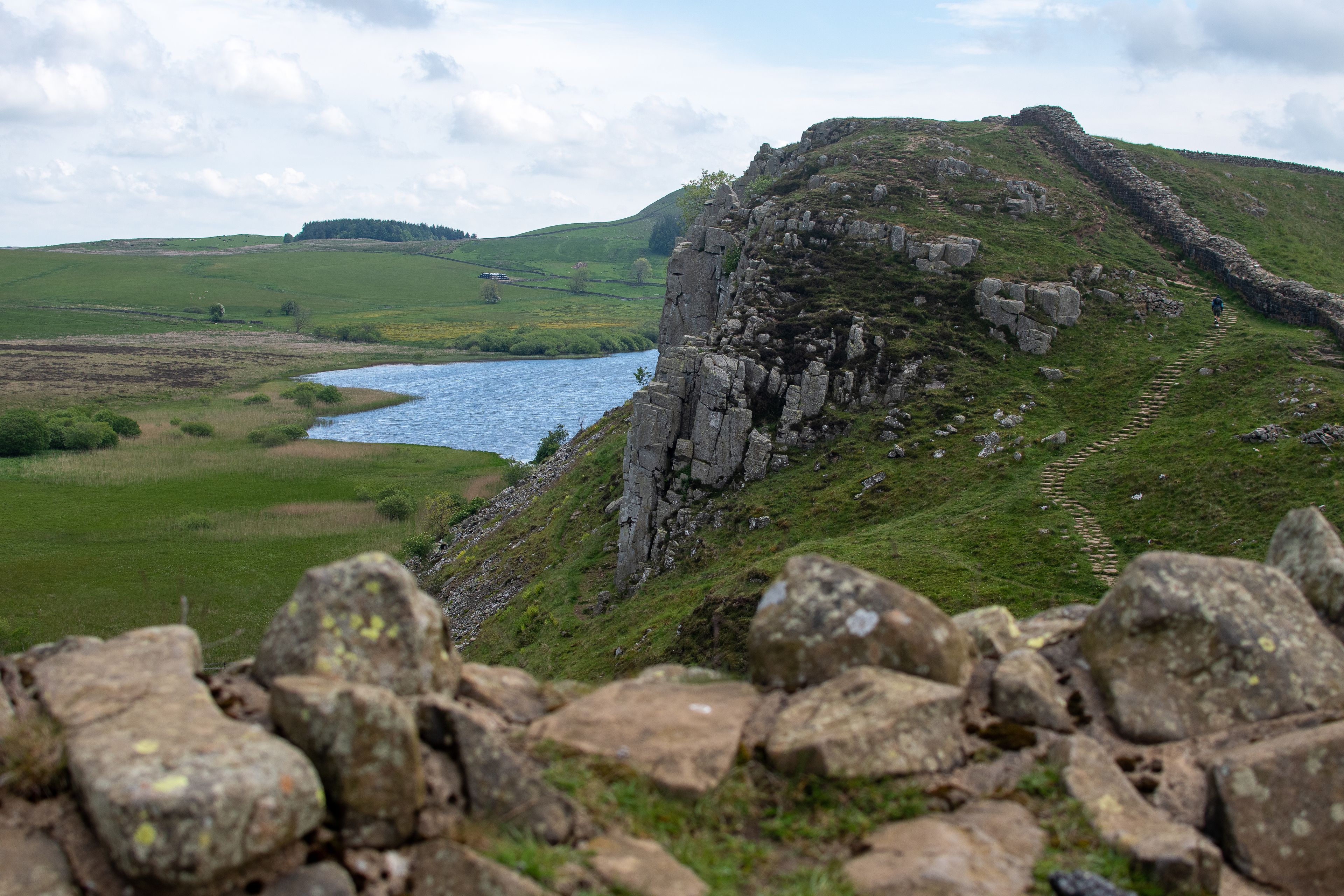 A rocky cliff with a river in the foreground, a grassy hill in the background, and a stone wall in the foreground.