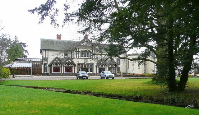 A large, historic building with a white facade and dark timber framing, surrounded by a well-maintained lawn and a large tree.