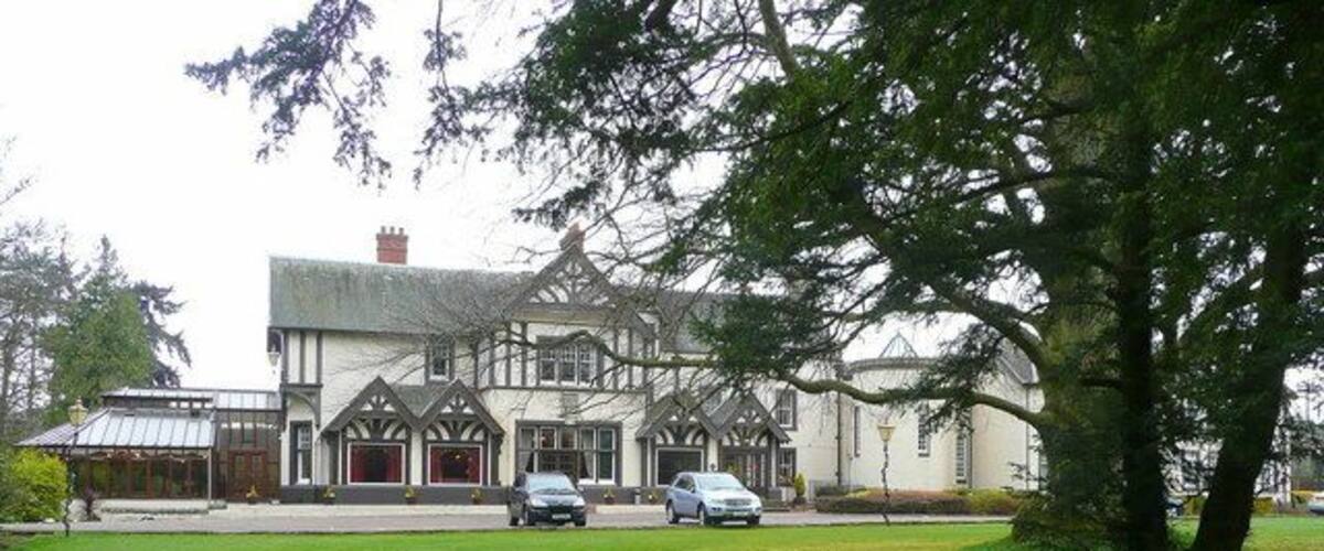 A large, historic building with a white facade and dark timber framing, surrounded by a well-maintained lawn and a large tree.