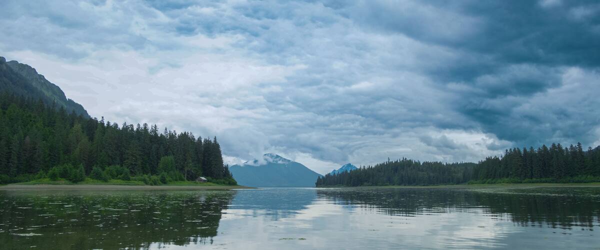A calm lake surrounded by dense forests and mountains under a cloudy sky.