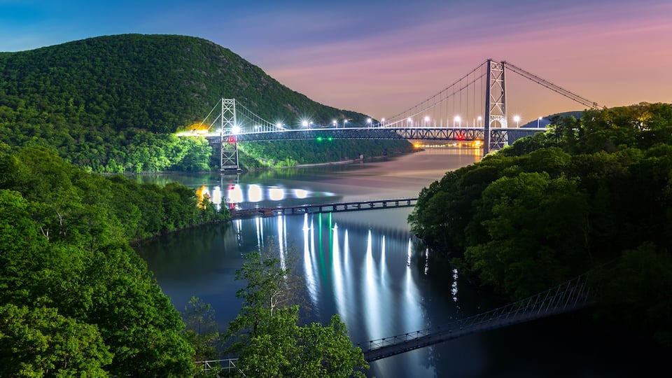 A suspension bridge over a calm river, surrounded by lush greenery and hills.