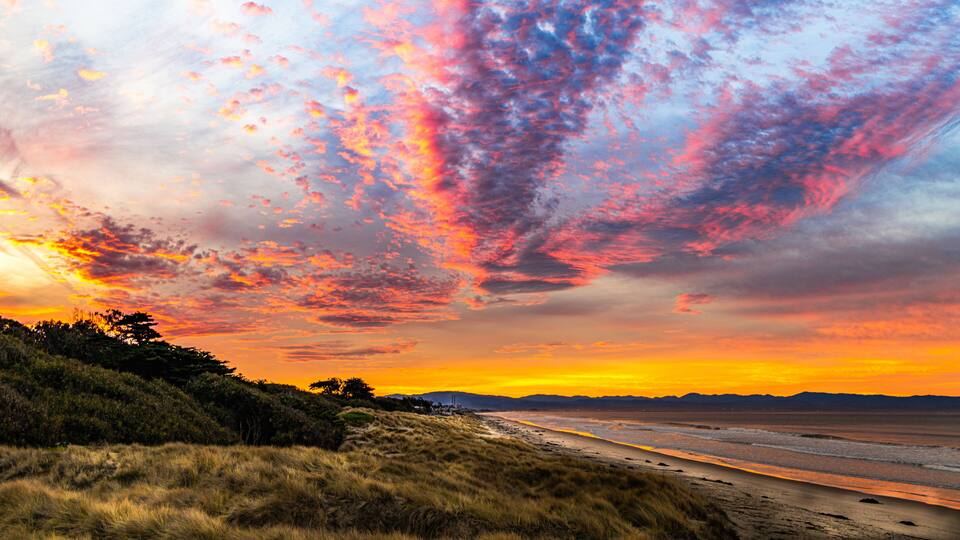 A sunset over a beach with a grassy foreground and distant mountains.