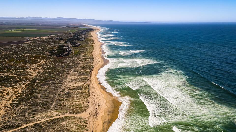 A coastal landscape with a sandy beach, ocean waves, and distant mountains.