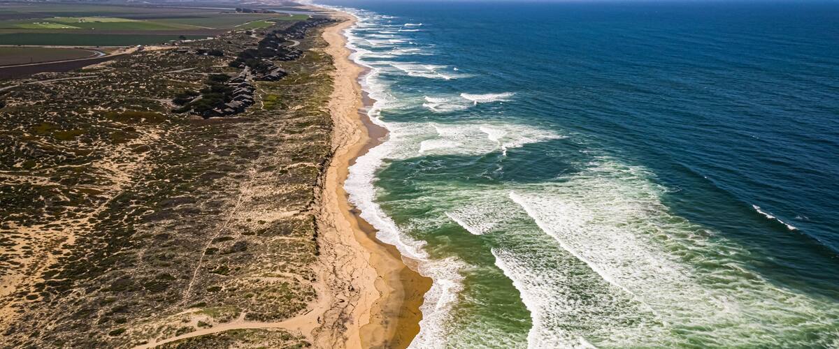 A coastal landscape with a sandy beach, ocean waves, and distant mountains.